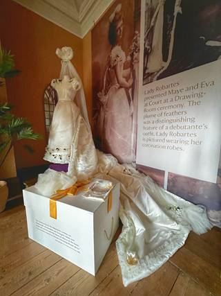 The replica gown on display at Lanhydrock. The gown is displayed on a plinth in the cenrtal background of the image with its full train spreading towards the central foreground. The veil and headress are suspended above the neckline of the gown. To the right of the gown there are floor to ceiling photographs of the original gown being worn by members of the Ager-Robartes family.