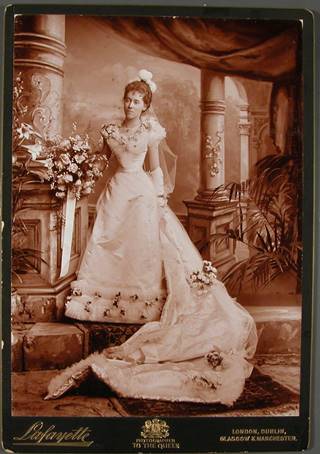 Everilda is standing on carpeted step with pillars and drapes behind, a fern on the right and wears a white dress with double frill at hem decorated with flowers, ostrich plumes in her hair, long gloves. She holds a large bouquet in her right hand which rests on a pillar. Her train which is arranged down the step also has flowers on it.