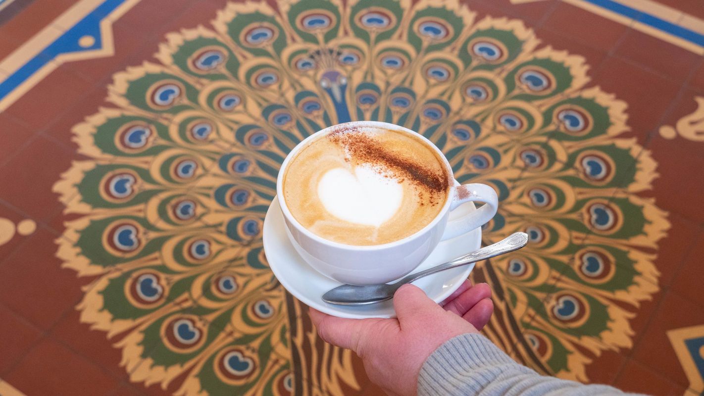 A hand holding a cup of milky coffee with a tiled floor depicting a peacock in the background.