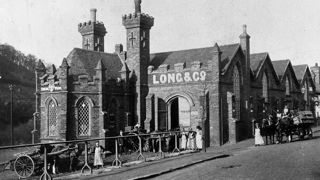 Black and white photograph showing brick building with crenellations and towers with horses and carriages and people in historic dress in the foreground.