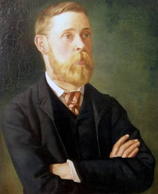 Portrait of a white man with short blond hair and a full blonde beard, wearing a black jacket and waistcoat, white shirt and striped tie.