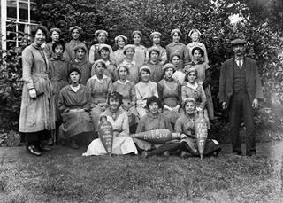 A formal group photograph of female munition workers in the First World War. Three girls at the front are holding bombs painted with anti-German slogans.
