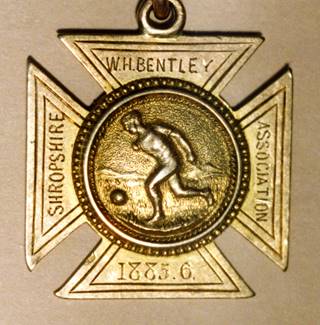Brass cross-shaped medal with footballer in centre dated 1885-6