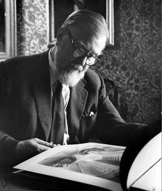 black and white photograph showing a man with a white beard and glasses sitting down. He is looking at a large book, and viewing a print in landscape. The print shows the interior of a train station. Behind him there are oil painting son the wall, and paisley patterned wallpaper.