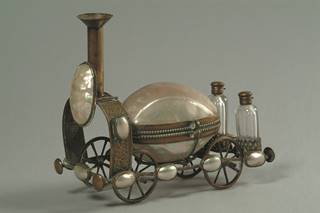 an object made to look like a 19th century train locomotive, but with the body made of a large pearl coloured shell fitted with brass. It sits on four brass wheels, and is decorated all around with smaller  pearl coloured shells. At the rear are two small glass bottles, with brass tops. Grey background.