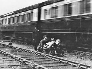 black and white photograph showing two figures knelling down by next to a railway track, while a third figure in railway uniform stands behind them. They are very close to a train moving past them, which is slightly blurry given its speed.