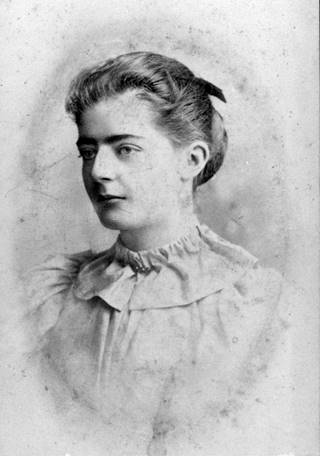 Black and white head and shoulder portrait photograph of a young woman. The photo has been taken half profile. He hair is in a bun and she is wearing a pale high necked blouse. Her face is relaxed but not smiling.