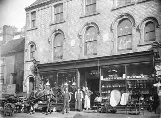Victorian ironmongers shop front with windows full of wares. three men stand outside the shop door, one is holding the reins of a horse who is pulling a waggon. More wares are displayed outside the shop front, including large tin baths and coal scuttles.