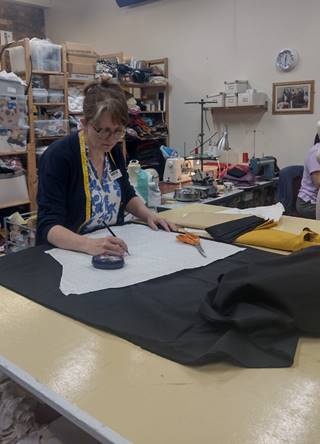 Alison is writing on a paper pattern piece which is laid out on top of a piece of black fabric that will be used for the replica costume. The fabric is laid across a dressmaker's cutting table, as well as a pair of fabric scissor and pins. In the background is the Cosutme Project workroom with shelves of fabric and haberdashery, and sewing machines.