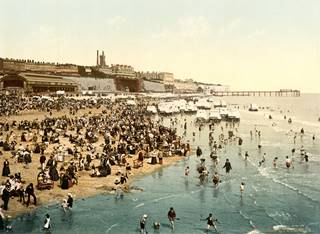 A very busy beach. The sand is almost entirely taken up with people sat on the beach, which is pictured left, whilst several dozen people play in the sea to the right of the images. In the distant background id the town of Ramsgate and its pier stretching into the sea.