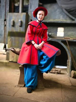 A person leans against an anvil in a Victorian woman's swimming costume. The cosutume consists of a wide brim red hat tied with a blue bow under the chin, a long red coat with bell shaped three-quarter length sleeves and an A-line skirt, and voluminous blue trousers that are tight at the ankle.
