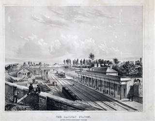 rAILWAY TRACKS RUN FROM THE BOTTOM RIGHT CORNER OF THE IMAGE INTO THE MIDDLE DISTANCE.  A small railway station building with a canopy is to the right of the tracks. In the foreground three boys sit on a bridge overlooking the railway tracks.