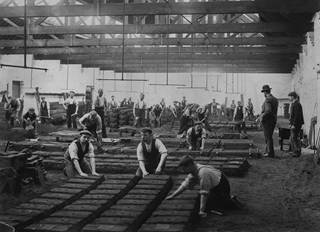 A balck and white photograph of the interior of a large industrial building. On the ground are rows of rectangular blocks of sand. Kneeling next to them and stood around them are workmen who are looking at the camera.