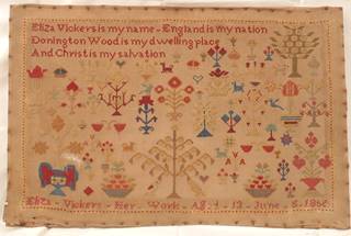 A needlework sampler done in cross stitch. The lettering is in red at the top left and along the bottom of the sampler. The rest of the design is of multiple plants and foliage in a mid-19th century cross stitch style in red, yellow, blue, orange and brown threads.