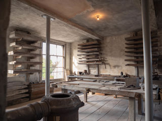 An old-looking empty room with wooden floorboards with wide close-placed shelves on the walls in the background and tables with tools on them in the foreground.