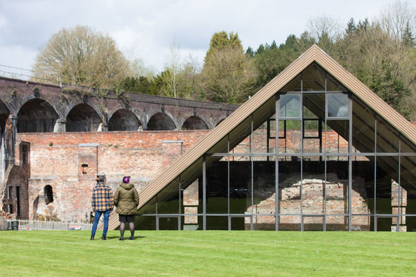 A man and a woman with their backs to the camera, standing on grass and looking at the Old Furnace, a glass-fronted triangular building.