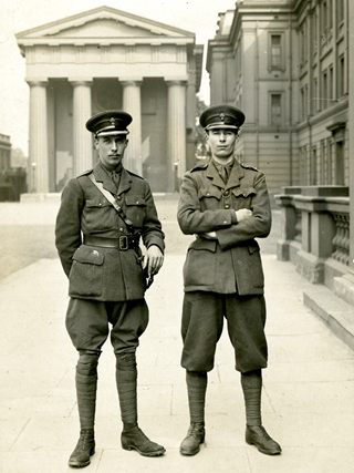 Two men in First World War army uniforms. Maurice, on the left, has one hand behind his back and one resting on his sword and is looking into the distance. The man next to him stares directly at the camera with his arms folded.