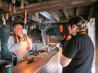 Two young women stand watching a costumed demonstrator, who is holding a lit candle, inside the candle factory at Blists Hill Victorian Town. The ceiling consists of criss crossing wooden beams, from which bunches of candle are dangling.