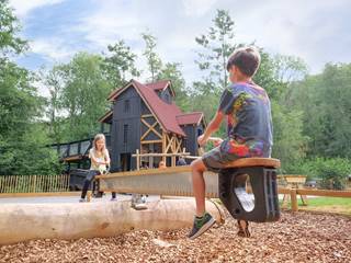 Two young children are sat on a large seesaw, styled as a ong saw. The backdrop consists of the main 'colliery' style building at Blists Hill Victorian Town's outdoor adventure, and it's surrounding woodland.