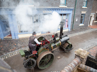 A man, dressed in Victorian costume, is driving a large green steam engine. The engine has two large rear wheels and one roller at the front. There is smoke coming out of a chimney stack on the front of the engine.
