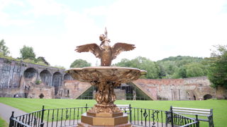 A gold fountain featuring a boy with his arms wrapped around a swan's neck in front of industrial brick buildings.
