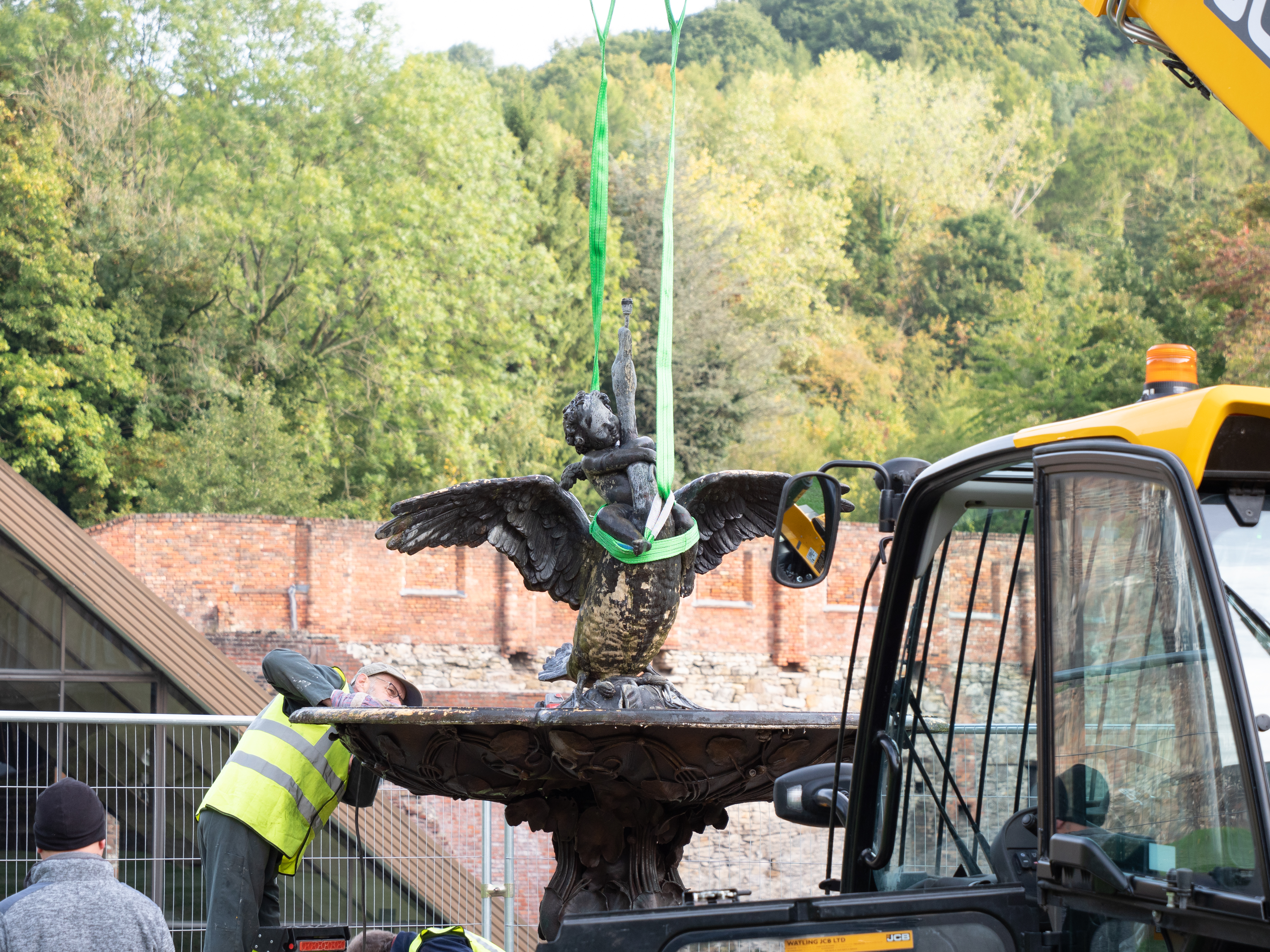 A blackened fountain featuring a boy with his arms wrapped around a swan's neck being lifted by a digger while a workman in a hi-vis jacket looks on.