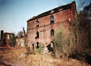 An old, slightly grainy photo of the blast furnaces at Blists Hill, made from brick and fallen into disrepair, with windows missing and vegetation growing over and around the buildings.