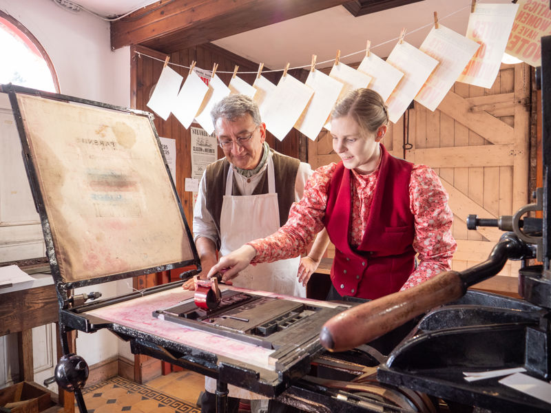 A smiling man and woman are stood at a Victorian printing press. They are dressed in Victorian costume and the woman is applying red ink to a print block. There are posters hanging behind them.